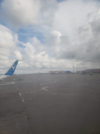 The image depicts an aerial view from an aircraft at Kazan International Airport in Stolbishche, Russia. The scene includes a portion of the aircraft's blue and green tail fin, adorned with a white logo, juxtaposed against a cloudy sky. Below, the airport tarmac is visible, marked with red lines and other painted markings. An individual in a lime green vest is standing on the tarmac. In the distance, the airport infrastructure is seen, including buildings, masts, and other airport equipment. The weather conditions appear overcast.