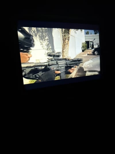 A person in a black tactical vest and black gloves is holding a black assault rifle with a scope. The person's face is partially visible, showing a determined expression. The rifle is pointed forward, suggesting readiness for action. The background depicts an urban environment with a white building, a tree with dappled sunlight, and a parked black car. The scene appears to be an outdoor setting, possibly a street or alleyway, in a city like Essen, Germany, given the architectural style and vegetation. The lighting suggests daytime. The overall impression is one of preparedness or a controlled situation involving the use of a firearm.