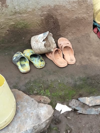 The scene depicts an outdoor ground-level area in Nyansiongo ward, Kenya. The ground is composed of dark, damp soil with patches of green moss. A cracked, textured earthen or plastered wall serves as the background. In the center, two pairs of worn plastic footwear are visible: one pair of mottled yellow/green and black clogs, and one pair of light brown open-toed sandals. Between the footwear lies a severely broken, light-colored plastic container. In the lower-left quadrant, a portion of a yellow plastic bucket rests on a large gray rock. Scattered debris, including dark fragments (possibly metal or plastic) and a piece of white paper, are present on the ground in the lower-right.