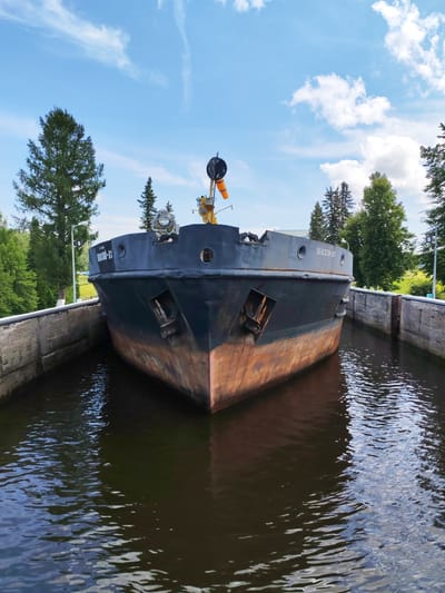 An eye-level, full-shot photograph captures a dark, rusted barge navigating through a canal lock on a clear day. The barge, painted a dark color with a prominent orange-brown rusted hull, dominates the frame, its bow pointed directly at the viewer. The canal walls, constructed of concrete and stone, flank the vessel, creating a narrow waterway. Lush green trees and tall pines line the canal banks, interspersed with a lamppost bearing a white number '5' sign. The sky above is a vibrant blue, dotted with fluffy white clouds. The water in the lock is dark and reflective, showing ripples and the distorted image of the sky and barge. The barge is inscribed with Russian lettering that reads "ШЕКСНИН-13" on its side. The scene appears to be located within or near the city of Belousovo, Russia, given the Cyrillic inscription on the barge, suggesting a possible connection to Russian waterways.