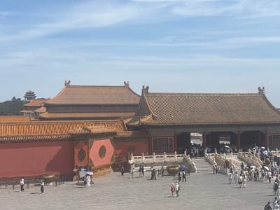 This media file depicts a wide-angle view of a historic courtyard within the Forbidden City in Beijing, China. The scene is bathed in bright daylight under a partly cloudy blue sky, suggesting it is daytime, likely late morning or afternoon.
The primary subjects are the imposing traditional Chinese architecture, characterized by their distinctive orange-tiled roofs with ornate decorations and the vibrant red walls of the buildings. The courtyard itself is paved with grey flagstones.
A significant number of people are present, appearing to be tourists, engaged in exploring the historical site. Many are walking around the courtyard, some are gathered in groups, and several are using umbrellas, possibly for shade from the sun. The general atmosphere seems to be one of sightseeing and mild tourism.
Notable details include large bronze urns placed strategically within the courtyard and elaborate stone railings along the staircases leading up to the buildings. In the background, a distant tower with a similar architectural style can be seen, hinting at the vastness of the complex. One sign with some illegible text is visible near the right side of the image. The overall impression is that of a bustling historical landmark on a clear, sunny day.
