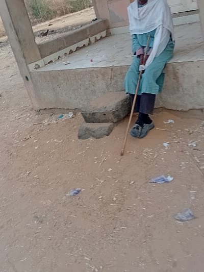 A person is captured in a sideways, mid-shot. Dressed in a white shirt and blue shorts, they hold a brown stick. The setting involves a corner of a building with visible steps and a concrete base. The ground is dirt, scattered with bits of trash, appearing earthy and dry.