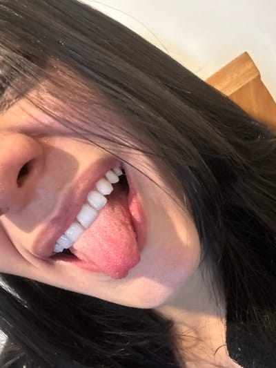 A close-up, angled photograph captures the lower portion of an individual's face. Dark, long hair frames the subject. The bridge and tip of a nose are visible on the upper left. The mouth is open, revealing a set of uniformly white upper teeth and a pink, textured tongue extended outwards. The immediate background consists of a plain light-colored surface, possibly a wall, with a small portion of a light brown, possibly wooden, object visible in the upper right corner. The lighting suggests an indoor environment. The specific location in the city of Bogota, Colombia, cannot be determined from the visual information provided in the image.