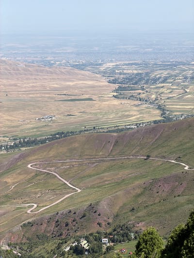 An aerial view captures a winding dirt road traversing a steep, grassy mountainside. The road snakes its way down the incline, forming multiple switchbacks. Below the mountain, a valley unfolds with patchwork fields and scattered trees. In the distance, a city sprawls across the plains, its buildings fading into a hazy horizon. The scene appears to be set in or around Kashka Suu, Kyrgyzstan, a region known for its mountainous landscapes and agricultural valleys. There are no discernible people or specific actions captured in the image.