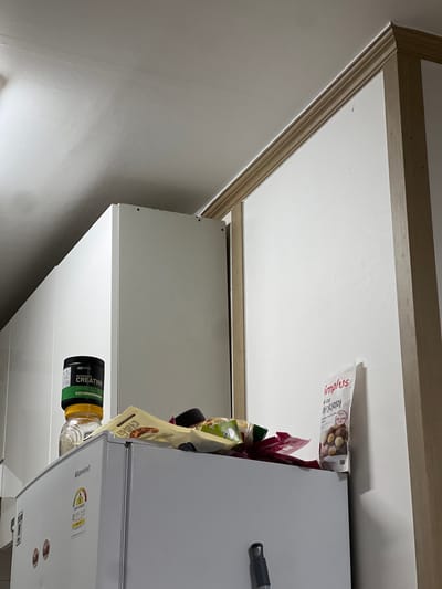 The image depicts the top of a white refrigerator in Geoje-si, South Korea, with various items placed on top. A black container labeled "MICRONIZED CREATINE" sits prominently. To its right, there is a clear glass jar with a yellow lid. Further to the right, there are several food packaging items, including a light beige bag with Korean text and an image of macadamia nuts, and a red pouch. The refrigerator has a "Klarwind" logo and energy efficiency rating stickers. The background consists of a white wall and a light-colored ceiling with crown molding.