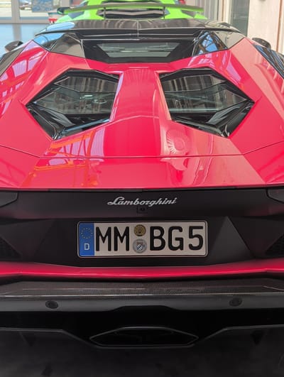 A close-up, rear view of a red Lamborghini is captured in an indoor setting, possibly a garage or showroom. The car's distinctive sharp lines and angular design are highlighted, with a prominent rear diffuser made of carbon fiber. A red LED strip runs horizontally across the rear of the car, adding a futuristic glow.

The license plate, mounted centrally, is a German plate with the characters "MM BG5". It also features the European Union stars and the abbreviation "D" for Deutschland. Below the main characters, there are circular emblems, one of which contains text that appears to say "BAYERN" and "STADT" with a depiction of a coat of arms. The word "Lamborghini" is subtly embossed on the black panel below the license plate.

The image is taken from a slightly elevated angle, looking down at the rear of the car. The lighting suggests an indoor environment, with reflections visible on the car's glossy red paintwork. The overall impression is one of luxury and high performance. The image is oriented vertically, with the top of the car at the left of the frame. There are no people visible in the image. The background is blurred but suggests a structured environment with some shelving or equipment. The location context of Böblingen, Germany, aligns with the German license plate and the style of the car.