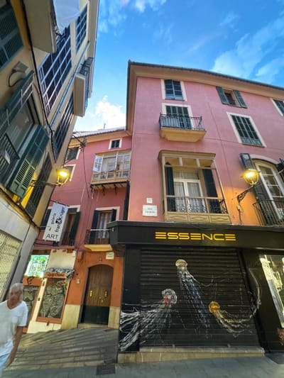This is a street-level, wide-angle photograph taken in Palma de Mallorca, Spain, looking up at buildings lining a narrow street. The buildings are a mix of architectural styles and colors, with the pink building dominating the right side of the frame and a yellow building on the left. The sky is blue with scattered clouds.
On the pink building, there are several windows with dark green shutters, some of which are open. A small balcony with black iron railings protrudes from one of the windows. Below this, a black metal shuttered storefront displays the illuminated yellow text "ESSENCE". The shutter itself is decorated with a mural of three jellyfish. Above this storefront, a narrow balcony with a wooden frame surrounds a window. Further up the pink building, another balcony is visible.
The yellow building on the left features numerous windows with dark green shutters, some of which are also open. A yellow streetlamp is attached to the side of this building, casting a warm light. A banner hangs from the yellow building, reading "galeria fran Krüger ART".
A portion of a man wearing a white t-shirt is visible on the left side of the image, walking away from the camera. The street itself is paved with cobblestones, and a few other details, such as a door and some graffiti on a lower level storefront, are also present. The overall impression is of a charming, narrow European street with a mix of residential and commercial spaces.