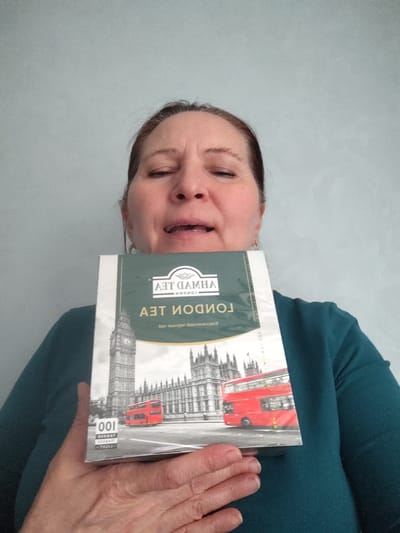A woman is depicted from the chest up, holding a box of Ahmad Tea London. Her face is positioned behind the upper portion of the box, with her eyes appearing closed or squinting, and her mouth slightly open. She is wearing a dark teal or green long-sleeved top. The background is a plain, light blue-grey wall, indicating an indoor setting.

The tea box is held upside down, making the text and imagery appear inverted. The front of the box features a prominent illustration of London landmarks, including Big Ben, the Houses of Parliament, and a red double-decker bus.

Visible text on the box, presented upside down, includes "AHMAD TEA" and "LONDON" in English. Below this, there is Russian text that reads "ЧАЙ ЛОНДОН" (Tea London) and "РУССКИЙ КЛАССИЧЕСКИЙ" (Russian Classic), suggesting it's a classic black tea. Near the bottom left, the number "100" is visible, along with the words "TAGGED TEABAGS."

The image captures a candid moment, possibly a blink or an unusual expression from the woman. The photograph was taken in Neftekamsk, Russia.