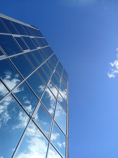A modern glass-facade building is viewed from a low angle, looking up towards the sky. The building's surface is composed of numerous rectangular glass panels, framed by metallic mullions, creating a grid-like pattern. The glass reflects the bright blue sky and scattered white clouds, giving the impression of the sky extending onto the building.
The scene is set outdoors, under a clear, sunny sky with a few wispy clouds. The time of day appears to be daytime, given the brightness of the sky and the reflections. There are no people visible in the image. The building's architecture suggests an urban or commercial setting, possibly an office building or a modern residential complex. The angle of the shot emphasizes the scale and geometry of the building against the expansive sky.
The reflections on the glass are a prominent feature, mirroring the surrounding environment and adding depth to the image. The colors are dominated by the vibrant blue of the sky and the muted tones of the building's structure and reflections. There is no visible text in the image. The overall impression is one of clean lines, modern design, and a harmonious interplay between the built environment and the natural sky.
