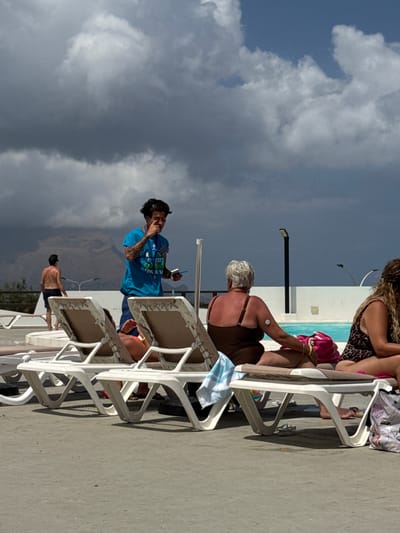 The image shows a group of people relaxing on lounge chairs by a swimming pool. The scene is outdoors, likely during the daytime, with a mix of bright sunlight and dark, stormy clouds in the sky. The people are dressed in swimwear and appear to be enjoying a leisurely day.
One person, a woman with grey hair, is sitting on a lounge chair wearing a brown swimsuit. She has a small medical device attached to her upper arm. Another person, a man wearing a blue t-shirt with text on it, is standing and appears to be talking on his phone. The t-shirt has text that reads "FINO AL PIÙ GRANDE" and "IL PIÙ GRANDE" in Italian, along with some other illegible text. A third person, a man with tanned skin, is seen from behind, wearing dark swim trunks. Other individuals are partially visible in the background, also seated on lounge chairs.
The setting appears to be a resort or a private pool area, with white plastic lounge chairs and a tiled pool edge visible. The ground is a concrete surface. The weather seems dynamic, with dramatic clouds suggesting a change in conditions. The overall mood is relaxed and casual, indicative of a vacation or a day spent by the water. The location is Capaci, Italy.