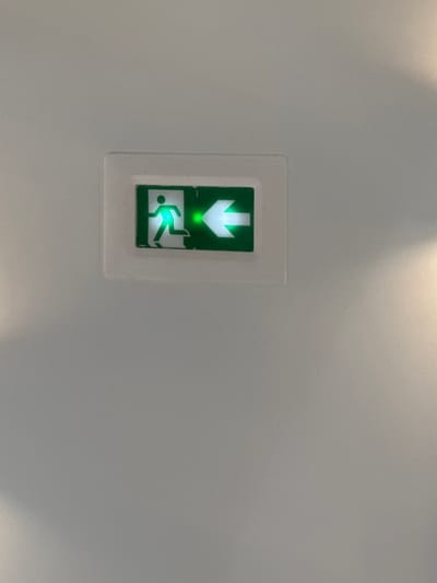 The image displays an illuminated green emergency exit sign mounted on a textured light grey wall. The sign is rectangular, set within a slightly raised, translucent or white plastic bezel. It features a pictogram of a running person facing left, followed by a green arrow pointing left, indicating the direction of egress. Both the running figure and the arrow are brightly illuminated green against a dark background within the sign's display area. The wall surface exhibits subtle variations in tone, with a brighter, warmer light source evident in the upper right portion of the frame, casting a soft glow. The scene is located in Orléans, France.