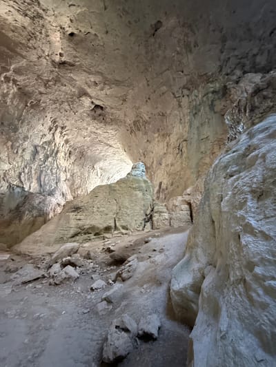 The image shows the interior of a cave, likely the Karlukovo Caves in Bulgaria. The cave features a large, cavernous space with rough, textured rock formations. The ceiling is high and arched, with visible cracks and irregularities in the rock. Natural light illuminates the cave from the far end, creating bright highlights and shadows on the surfaces. The floor of the cave is covered in dirt and loose rocks, with a path winding through the formations. Several large rock pillars and boulders are present, some appearing to be stalagmites or other mineral deposits. The overall impression is one of a vast, ancient underground space.