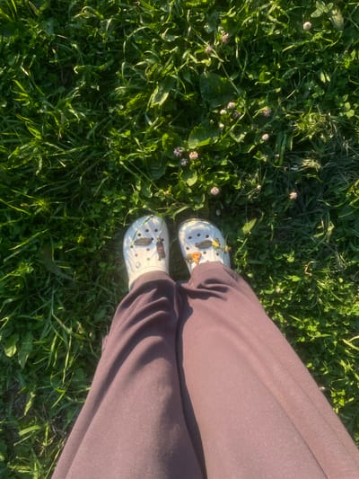 The image shows a person's legs and feet, wearing white Crocs with charms, standing on a patch of green grass. The Crocs have charms including a slice of pizza and a Coca-Cola bottle. The person is wearing dark purple sweatpants. The grass is lush and green, with some small pink clover flowers interspersed. The scene is illuminated by sunlight, creating shadows and highlighting the texture of the grass and fabric. The image is taken from a top-down perspective. The location is identified as Podshivalovo, Russia.