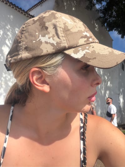 The image shows a close-up profile view of a young woman wearing a camouflage baseball cap and a patterned bikini top. She has blonde hair pulled back in a ponytail and is looking to her right. Her skin is illuminated by sunlight, casting shadows on her face and shoulder. In the background, a white building with a tiled roof is visible, along with trees and a man wearing sunglasses and a white t-shirt. The scene appears to be outdoors in a sunny location. Based on visual cues, this location could be Icod De Los Vinos, Spain, a town known for its sunny climate and white-washed architecture.