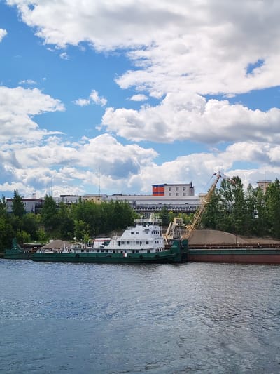 This image depicts a wide, calm river in Yaroslavl, Russia, under a bright blue sky with fluffy white clouds. In the foreground, the blue water shows ripples. Moored along the right bank are two large vessels. The vessel on the left is a green and white ship with a crane. The name "КОНСТАНТИН МАСЛАКОВ" is visible on its side. This ship appears to be loaded with loose material. To its right is a long barge, also filled with what appears to be gravel or sand. A tall yellow crane, likely for loading and unloading, extends from the barge. In the background, beyond a line of lush green trees, are several industrial buildings and structures, suggesting an industrial or port area of the city. The scene is tranquil, showcasing a typical day at a riverside industrial location.