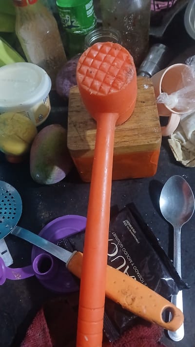 The image presents a cluttered scene on a dark surface. Visible objects include a meat tenderizer (orange), a wooden block, a metal spoon, a spatula (orange handle), a coffee packet ("AUCA"), several glass bottles, a plastic container, a mango, purple plastic kitchen utensils, and a red cloth. No people or actions are discernible in the image. There's no specific indication of location in Putucual, Venezuela.
