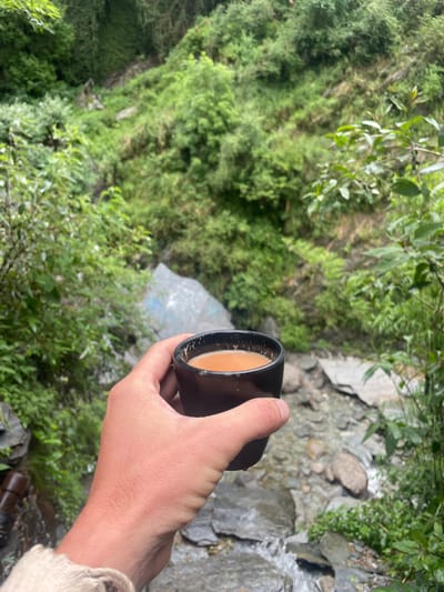 In the city of Bhagsu, India, a hand holds a black ceramic mug filled with a light brown beverage, likely tea, in the foreground. The mug has a slightly worn rim with white specks. The hand is pale and rests against a light-colored, loosely woven fabric. Behind the hand and mug, a rocky stream flows with clear water. Lush green vegetation covers the surrounding hillside, with trees and foliage creating a dense, natural backdrop. The scene appears to be outdoors, possibly on a hiking trail or near a waterfall, given the natural environment and the presence of water. The lighting suggests it is daytime.