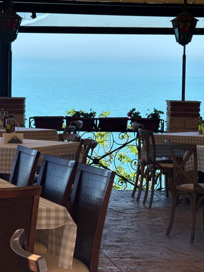 A photograph taken at an angle shows an outdoor dining area overlooking a body of water. The scene is likely a restaurant terrace in the city of Balgarevo, Bulgaria. The space is furnished with wooden tables and chairs covered in checkered tablecloths. Decorative wrought iron railings line the edge of the terrace, adorned with potted plants containing greenery and red flowers. Beyond the railing, the calm, light blue water stretches out to the horizon, meeting a clear sky. A decorative lantern hangs from the ceiling of the terrace, partially visible in the upper left corner of the image. Several chairs are stacked at an angle to the terrace floor.