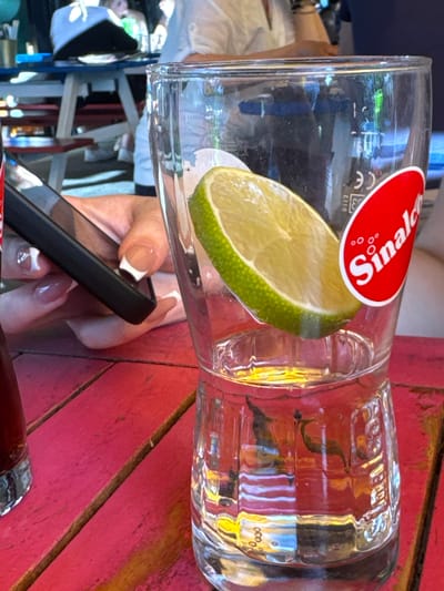 A person's hands are visible, holding a black smartphone. The phone is positioned in front of a glass that has a lime wedge inside it and a "Sinalco" logo on the outside. The glass is sitting on a red wooden table. In the background, other people are seated at a table with blue and white tablecloths. The scene appears to be outdoors, possibly at a beer garden or restaurant.
