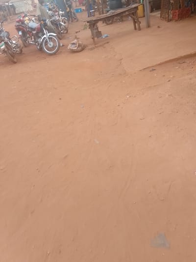 The image depicts a dirt road or open area in Ejule, Nigeria. Several motorcycles are parked on the left side. A wooden table or structure stands in the upper center. Some people are standing near the motorcycles and the table. The ground is dry and uneven. There are some objects and containers in the background.
