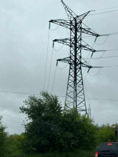 An outdoor, daytime, medium shot captures a towering electrical pylon against an overcast sky, as viewed from a vehicle. The pylon, constructed of dark metal lattice, dominates the upper right quadrant of the frame, with power lines extending from its crossbars. To the lower left of the pylon, lush green trees and foliage fill the mid-ground. In the bottom right corner, the rear of a dark-colored car with its brake lights illuminated is visible. The scene appears to be located in an area with natural vegetation, possibly on the outskirts of Krasnokamsk, Russia, indicated by the presence of the infrastructure and the surrounding greenery.