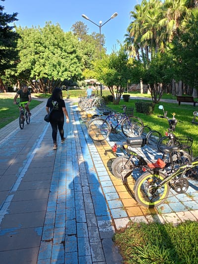In Antalya, Turkey, a sunny day in a park setting shows a paved walkway with a blue and yellow painted pattern. A man wearing a dark green t-shirt and neon green shorts cycles on the left side of the path, casting a shadow. A woman in a black t-shirt and black leggings walks away from the camera on the same path, with a black bag slung over her shoulder. Several bicycles are parked in a rack on the right side of the path, with some leaning against the rack. One electric bike, black and green, is prominent in the foreground. Palm trees and lush green trees line the background, with a park bench visible in the far right. A streetlamp stands tall on the left side of the walkway. The ground is a mix of paved tiles and grassy areas.