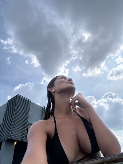 This low-angle photograph captures a person looking upwards against a dramatic sky, set in the urban environment of Ampang Jaya Municipal Council, Malaysia. The main subject is a female, seen from the chest up, wearing a black bikini or swimsuit top. Her skin and wet dark hair are adorned with numerous water droplets, indicating she has recently been in or near water, possibly a pool. Her head is tilted back and her gaze is directed towards the sky, with one hand gently touching her neck or collarbone area.
The sky occupies the majority of the frame, showcasing a mix of bright white, sunlit clouds and darker grey clouds, suggesting a partly cloudy but bright daytime. Patches of lighter blue sky are visible between the cloud formations. On the right side of the image, a segment of a modern building with a distinctive dark, slatted or ribbed facade rises sharply, providing an architectural element to the scene. The perspective makes the building appear tall and imposing. The overall impression is one of serene contemplation or relaxation in an outdoor, possibly poolside or rooftop setting, under a vast and dynamic sky. There is no visible text in the image.