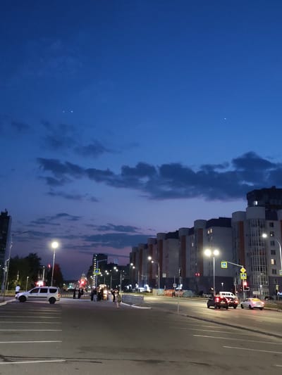 This is a vertical, eye-level, nighttime photograph of a city street in Nizhnevartovsk, Russia. The sky is a deep blue with wisps of clouds and two small, bright celestial objects are visible. The street is paved, with multiple lanes and white lane markings. On the right side of the street, a modern apartment building complex with balconies and lit windows stretches into the distance. Streetlights illuminate the scene.
Several cars are present: a silver minivan is parked in the foreground on the left, and a white car and two dark-colored cars are driving in the middle ground. Pedestrians are visible on the sidewalk and near a crosswalk. Traffic lights and street signs, including pedestrian crossing signs, are posted. In the distance, more buildings and streetlights are visible. The overall atmosphere is that of a typical urban street scene at dusk or after dark.