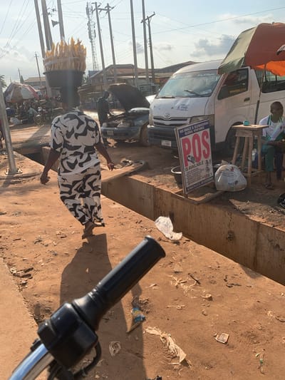 This media file captures a street scene in Shagamu, Nigeria, bathed in the warm glow of what appears to be late afternoon or early morning sun, casting long shadows. The foreground is dominated by the handlebars of a vehicle, likely a motorcycle, indicating the perspective of someone on or near it.
The scene depicts a bustling roadside environment. A white minibus, possibly a public transport vehicle, is parked, with visible signage on its side. A prominent sign advertises "POS IS AVAILABLE" for "HIS GRACE & OLUWABUNMI GLOBAL VENTURES," listing services such as cash withdrawal, money transfer, bill payment, and recharge cards, along with contact phone numbers. There are also bags and debris scattered on the dusty ground, suggesting a busy commercial area.
A woman walks away from the camera, wearing a black and white patterned dress. To her left, a vendor displays a large bundle of corn on the cob, wrapped and ready for sale, atop a dark container. In the background, other vehicles, including cars and motorcycles, are visible, along with utility poles and wires, typical of an urban Nigerian setting. The overall impression is one of everyday commerce and activity on a Nigerian street.