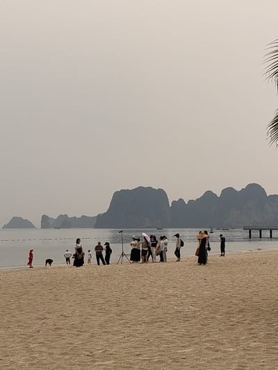 This image depicts a tranquil beach scene in Ha Long Ward, Vietnam, under an overcast sky.
In the foreground, a wide expanse of light-colored sand stretches across the bottom portion of the frame. A group of approximately 15-20 individuals is gathered near the water's edge, forming the primary subjects of the image. Many are dressed in casual clothing, with some wearing hats and one person holding an open umbrella. Their arrangement suggests a coordinated activity, possibly a photoshoot, as a camera on a tripod is prominently visible on the right side of the group. One person in a striking red outfit stands on the far left of the group, near another individual kneeling close to the water.
The midground features calm, grey-toned ocean water, dotted with a line of buoys or markers. Several small boats are scattered in the distance across the water. A wooden pier with support pilings extends into the sea on the right side of the frame.
The background is dominated by the distinctive, dark silhouettes of karst limestone mountains or islands, characteristic of Ha Long Bay. These geological formations are partially veiled by atmospheric haze, blending into the light grey, uniform sky. A section of a palm frond is visible along the far right edge of the image.
The overall atmosphere is quiet and subdued due to the overcast conditions, which create soft, diffused lighting. The primary activity appears to be a photography session or a group gathering, possibly for commemorative pictures, set against the iconic Ha Long Bay landscape. No visible text is discernible in the image.