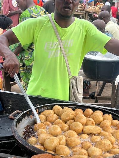 The image shows a man in Egan, Nigeria, preparing fried dough balls in a large, black wok filled with hot oil. The man is wearing a bright green t-shirt and holding a large, metal spoon. He appears to be flipping or stirring the dough balls in the oil. A small, white shoulder bag is visible near the man's waist. The surrounding environment suggests a busy, outdoor market or street food setting with other people nearby.