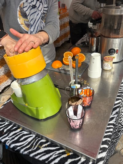 An indoor scene in Blois, France, depicts a commercial food preparation area, likely a market stall or cafe, focused on beverage service. In the foreground, a person's hands are actively pressing a halved orange onto the yellow reamer of a lime green electric juicer. A white paper cup is positioned to collect the juice from the juicer's spout. The person wears a gold ring on their left ring finger and a smartwatch on their left wrist, and is dressed in a grey long-sleeved top with sequin embellishment and a striped scarf.

On the stainless steel countertop to the right of the juicer, several whole and halved oranges rest on a dark, rectangular cutting surface, alongside two clear plastic utensils. Further right, a stack of white paper cups, a small jar labeled "Les Ruchers," and a silver commercial beverage dispenser, possibly for hot chocolate, are visible. A small metal pitcher with a stirring spoon is situated near the dispenser. Scattered coins are on the counter surface. Three small glasses contain various packets, including sugar and stirrers. The countertop is covered underneath by a black and white zebra-print fabric. In the background, partially obscured, another individual in a grey hoodie and dark trousers operates another station. The environment suggests a functional, semi-public setting.
