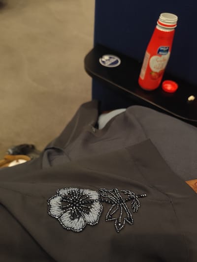 The image captures a close-up of a dark grey garment, likely a thobe or abaya, adorned with intricate beaded embroidery. The embroidery, positioned prominently in the foreground, consists of a stylized flower crafted with light blue and dark grey beads, and an accompanying leaf motif made from dark grey beads.
In the blurred background, a small black tray table is visible. On the tray sits a tall, red plastic bottle, resembling a juice bottle, with its white-striped cap removed and placed next to it. The bottle's label features a prominent blue and white "Almarai" logo, along with Arabic text that clearly indicates "شراب الفراولة" (Strawberry Drink). A few small, white specks, possibly remnants of a snack, are also scattered on the tray. The tray table itself has a circular, silver-colored emblem. Behind the tray is a dark blue surface, suggesting upholstered seating. The floor or a similar flat surface in the foreground is a light, blurred beige.
The scene suggests an indoor setting, likely within a mode of transport such as an airplane or train, given the presence of a tray table and seat design. There is no direct activity visible; the image presents a still life, hinting at a moment of rest or travel. The notable beaded garment, combined with the location context of Jeddah, Saudi Arabia, suggests a cultural significance to the attire. No specific time of day or emotions are discernible.