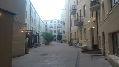 The image depicts an outdoor courtyard of a building complex in Łódź, Poland. The courtyard is paved with brickwork and lined on both sides by multi-story buildings. The architecture suggests a modern, possibly residential or commercial development with balconies and large windows. String lights are visible, hanging across the courtyard, some illuminated. Several potted plants and trees are placed strategically throughout the space. In the foreground, a person is seated on a chair on the left side, and on the right, there are two bicycles leaning against a railing. Further into the courtyard, more chairs are set up, and a food delivery person with a yellow backpack is visible on a bicycle. The overall atmosphere is calm and serene, with a hint of a commercial or hospitality setting given the presence of outdoor seating and the food delivery person.