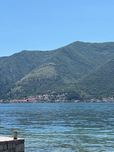 The image displays a panoramic view of Dobrota, Montenegro, under a clear blue sky. In the foreground, a stone pier or wall, constructed from light-colored, rough-hewn stones, extends from the lower-left corner into the water. A cylindrical stone post rests on this structure. The water in the midground is a deep blue-green, exhibiting ripples and reflections, with lighter shades closer to the foreground structure suggesting shallower depth. Across the expanse of water, the town of Dobrota is visible along the shoreline. It comprises numerous buildings characterized by terracotta or red-tiled roofs and light-colored walls, clustered along the waterfront and ascending the lower slopes of the mountains. Several small, white specks, indicative of boats, are present on the water near the town. Dominating the background are steep, verdant mountains, densely covered with green trees and vegetation. A prominent slender communication tower is positioned on the summit of one of the mountains in the upper-left quadrant. The sky is uniformly clear and bright blue, occupying the top third of the frame. No individuals are discernible within the image.