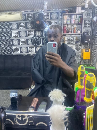 An individual is captured in a mirror selfie inside a barbershop in Samaru, Nigeria. The subject is a young Black male, appearing to be in his 20s or early 30s, with a close-cut fade hairstyle and lightened or bleached hair on the top of his head. He is wearing a black barber's cape. His right hand holds a grey smartphone with a red camera module, positioned to take the photo. A gold-toned wristwatch is visible on his left wrist.
The background, reflected in the mirror, reveals the barbershop's interior. The walls are covered in two distinct wallpapers: the left side features a black and white pattern with interlocking "Gucci" logos, while the right side displays a black and white geometric Greek key or labyrinth pattern. The ceiling above appears to be corrugated. A white wall-mounted air conditioning unit is positioned on the upper left, and a black wall-mounted oscillating fan is on the upper right. A black circular wall clock, reflected to show approximately 10:45, is visible below the fan. A black upholstered sofa or bench is against the left wall beneath the "Gucci" wallpaper.
A white wall-mounted shelf unit, stocked with various barber products in containers and bottles, is situated on the wall behind the subject's right shoulder (left in reflection). To the right of these shelves, a yellow electrical box labeled "TABA," possibly an inverter or stabilizer, is mounted on the wall.
The foreground, on the counter directly below the mirror, displays an arrangement of barber tools. These include a black hair trimmer with a textured grip, a rose gold metallic trimmer, and a grey trimmer, all resting on a dark stand. Several upright brushes with soft bristles, both white and black neck dusters, are visible. Additionally, yellow-handled brushes and combs, some with noticeable wear and chipped paint, are held within purple and green organizing containers.