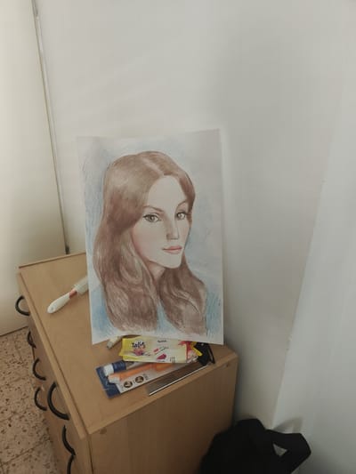 A colored pencil drawing of a woman's face is positioned on top of a light-brown, three-drawer dresser. The drawing depicts a woman with brown hair and light skin, looking to her right. The background of the drawing is a light blue. Beside the drawing, on the dresser, are a white hairbrush, a small packet of wet wipes with the brand name "Zalia" visible, and a packaged toothbrush and toothpaste set. The dresser is situated in a room with a tiled floor and white walls. A black bag is visible in the lower right corner of the image, partially obscuring the wall. The lighting in the image suggests an indoor setting, possibly a bedroom or a study area. No specific landmarks in Netanya, Israel, are identifiable.