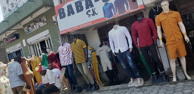 The image shows the exterior of a clothing store in Rubavu, Rwanda, located at latitude -1.668 and longitude 29.268. The store is called "BABA SK," with a sign above the entrance indicating a contact number. The store's facade is painted in a light green color, with a white awning above the entrance. The storefront is filled with mannequins wearing various clothing items, including a white shirt and jeans, a red button-down shirt with black pants, a yellow and black striped shirt with grey pants, a white t-shirt with beige shorts, a green shirt, and a red and black dress. There is a sign above the store with the text "HOUSE OF THE KOPA COLLECTION" which is likely a brand of clothing sold in the store. Two men stand in front of the shop, the one on the left is wearing a grey t-shirt and grey pants, and the one on the right is wearing a white shirt and jeans. There is a wooden stool in front of them. The ground in front of the store is made of grey concrete paving stones. The image is taken during the day with bright sunlight.  The focus of the image is on the clothing store and the mannequins displaying the clothes.