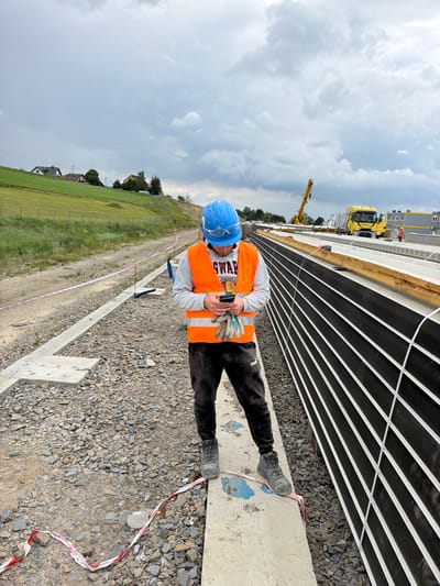 In Męcina, Poland, a construction worker wearing a blue hard hat, safety glasses, and an orange safety vest stands on a concrete beam, looking at his mobile phone. He is dressed in dark pants and work boots, with a pair of work gloves in his left hand. The worker is surrounded by a construction site with gravel on the ground and what appears to be modular building components or large structural elements stacked nearby. In the background, a yellow crane and a yellow truck are visible, along with a line of prefabricated buildings. The sky is overcast with scattered clouds, and a grassy hillside with houses can be seen in the distance. Red and white caution tape is present on the ground around the worker.
