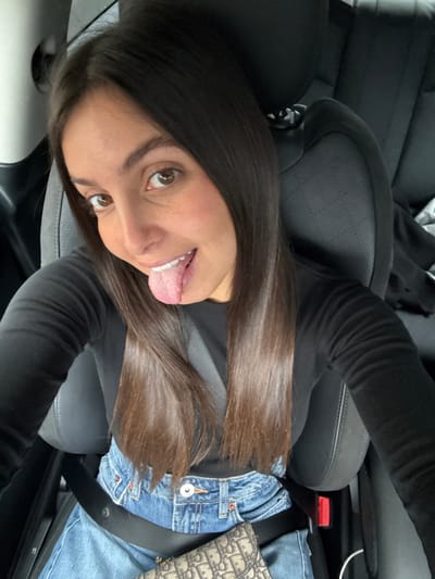 A female subject with long, straight dark brown hair and brown eyes is depicted from the chest up, seated inside a vehicle. She wears a black long-sleeved top, light blue denim jeans, and a light-colored belt bag with a repeating dark logo pattern. A black seatbelt is engaged across her torso and lap. The subject faces the camera directly, smiling and extending her tongue. The vehicle interior features dark-toned, possibly leather, seats; a quilted pattern is visible on the backrest of the seat behind her. A light-colored overhead lining or pillar is visible in the upper left of the frame. A white cable is present near the seatbelt buckle on the right. No visual identifiers within the image confirm a specific location in Antibes, France; the scene depicts a generic car interior.