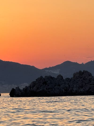 The image depicts a serene coastal scene at sunset. The sky is a gradient of warm orange and yellow hues, indicative of the golden hour. In the foreground, the textured surface of the sea stretches across the right side of the frame, reflecting the soft light of the setting sun. The water's surface is marked by ripples, creating a dynamic interplay of light and shadow.

To the left of the water, a dark, rugged cliff face dominates the middle ground. Its rough texture and deep shadows suggest a rocky, mountainous terrain. Behind the cliff, a silhouette of a distant mountain range is visible against the sunset sky. The outline of the mountains is softened by the atmospheric haze of twilight.

The scene is devoid of any prominent human figures or man-made objects, emphasizing the natural beauty of the location. The overall mood is peaceful and tranquil, evoking a sense of calm and reflection. The time of day is clearly sunset, characterized by the warm, diffused light. The location is identifiable as Sveti Stefan, Montenegro, known for its picturesque coastline and dramatic cliffs. There is no visible text in the image.