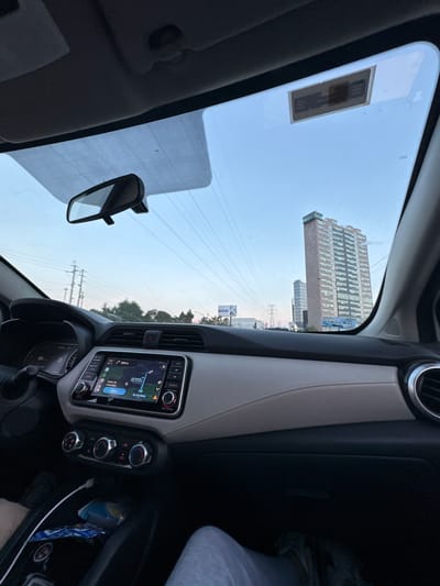 The view is from inside a car, likely a Nissan Kicks, driving along Avenida Las Torres in Heroica Puebla de Zaragoza, Mexico. Through the windshield, there is a tall building, probably the Torres JV III. Power lines stretch across the sky. The rear-view mirror is visible. The dashboard features a navigation system showing a map, and climate control knobs.