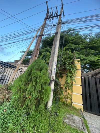 The image depicts a street scene in Ilado, Nigeria. The foreground shows overgrown vegetation, including tall grass and a shrub. A fallen concrete utility pole lies horizontally, with multiple attached power and communication cables. The pole is marked with the designation "FOR-IL-0012". Behind the fallen pole, a yellow brick wall with a dark metal gate is visible. Further back, additional trees and vegetation can be seen. Another utility pole stands upright, also with numerous cables attached. The sky is partially visible, suggesting either dawn or dusk. No people or animals are visible in the frame.