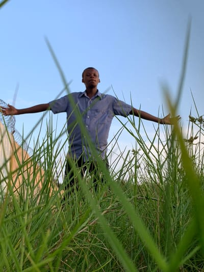 A young man, likely a teenager or young adult, stands amidst tall, vibrant green grass in an outdoor setting. He is wearing a light blue, striped short-sleeved collared shirt and dark trousers. He faces the camera directly, displaying a calm or slightly smiling expression. His right arm is raised and extended upwards, with his hand open, while his left arm is down by his side.

The immediate foreground and midground are filled with dense, green grass, which appears slightly out of focus, giving a sense of depth and a low-angle perspective, as if the photo was taken from within the grass. Behind the man, a clear, light blue sky dominates the background, indicating good weather conditions. A blurry, light-colored structure, possibly a fence or part of a building, is faintly visible in the upper left background, partially obscured by the grass and the sky.

The scene depicts the man in a static pose, possibly reaching, waving, or simply posing for the photograph in a natural environment. The bright, even lighting suggests it is daytime, likely late morning or afternoon, without harsh shadows. The image conveys a serene outdoor moment in a grassy area, consistent with a natural landscape in Ibasa, Nigeria. There is no visible text.