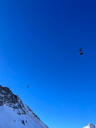 The image presents a striking view of a vibrant, clear blue sky dominating the upper and left portions of the frame, with a snow-covered mountain and rocky cliff face occupying the right side. Several parallel cable car lines stretch diagonally across the bright sky.

Two cable car cabins are visible on these lines. A larger, dark-colored cabin with prominent white components is positioned in the upper left quadrant, appearing to move along the cables. A second, smaller cabin can be seen further along the lines towards the mid-right, closer to the mountain's edge. A faint, thin white contrail, likely from a distant aircraft, is discernible in the upper right part of the sky.

The scene is set in a high-altitude mountain environment, characteristic of Courmayeur, Italy. It depicts a bright, sunny day with no clouds, suggesting excellent weather conditions. The strong sunlight creates clear visibility and highlights the snow and rock textures on the mountain. The perspective is from a low angle, looking upwards, emphasizing the vastness of the sky and the scale of the cable car system against the mountain backdrop. No visible text is present in the image.
