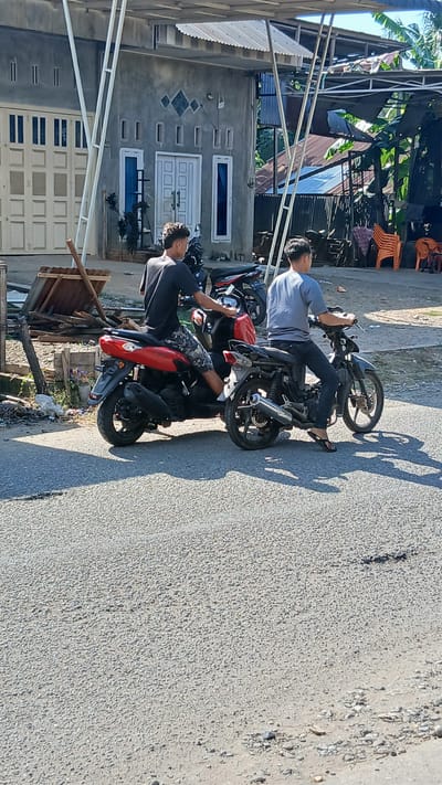 In Keudejungkagajah, Indonesia, two young men are seen from behind, each sitting on a motorcycle on a paved road. The man on the left, wearing a black t-shirt and camouflage shorts, is on a red motorcycle. The man on the right, dressed in a gray t-shirt and dark pants, is on a black motorcycle. They appear to be facing forward, possibly about to start riding. Behind them, a grey building with white-framed doors and windows is visible, along with some wooden planks and other debris. The environment suggests a rural or suburban setting with tropical vegetation visible in the background under a clear sky.