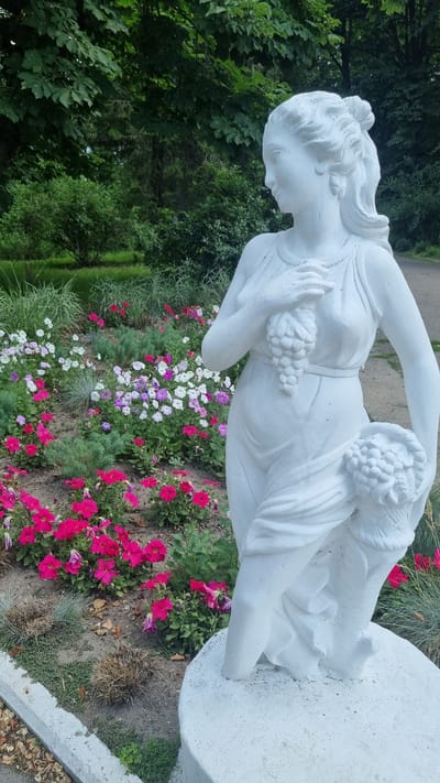 A white stone statue of a woman holding grapes and a basket of fruit stands in a flower bed in Smila, Ukraine. The statue is life-sized and depicts a classical female figure with flowing drapery. She is positioned to the right of the frame, with her gaze directed to the left. The flower bed in front of the statue contains a vibrant mix of pink, white, and purple petunias, interspersed with some greenery and dried foliage. Behind the statue and flower bed, a pathway is visible, bordered by lush green trees and bushes, suggesting a park or garden setting. The overall scene is serene and picturesque, with the white statue providing a striking contrast against the colorful flowers and natural backdrop.