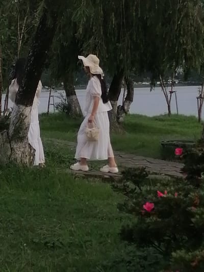 The image shows two individuals in white dresses standing on a stone pathway in a grassy area with trees and a body of water in the background. The person in the foreground is wearing a wide-brimmed hat and carrying a small woven bag. The person in the background is partially obscured by a tree trunk. The scene appears to be a park or garden setting. The location is specified as Lam Vien - Da Lat, Vietnam.