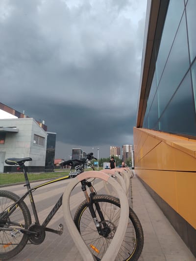 The image depicts a bicycle rack with two bicycles parked in front of a modern building with a glass facade and orange trim. The sky is heavily overcast with dark, ominous clouds, suggesting impending rain or a storm. In the background, several apartment buildings are visible, characteristic of urban development. A few people are walking near the building and in the distance. The scene is likely in the city of Kommunarka, Russia, given the style of architecture and residential buildings. The foreground is dominated by the bicycles and the bike rack, with the building and sky filling the rest of the frame.