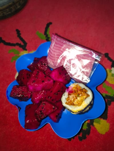 A blue, flower-shaped plastic plate is centrally positioned on a red textile surface with a black, green, and yellow abstract or floral pattern. The plate contains several items of food. To the left, numerous irregular chunks of cut red-fleshed dragon fruit with visible black seeds are arranged. To the lower right, a single piece of a sushi-like roll is visible, featuring a white rice exterior, a dark seaweed wrap, and an interior filling of what appears to be cooked chicken or a similar protein with other ingredients and a sauce. A small pink/purple topping is on the roll. In the upper right quadrant of the plate, a small, clear plastic bag contains thin, pink-colored strips of food. A dark, circular object is partially visible in the upper left background. The scene is located in An Khe Ward, Vietnam.
